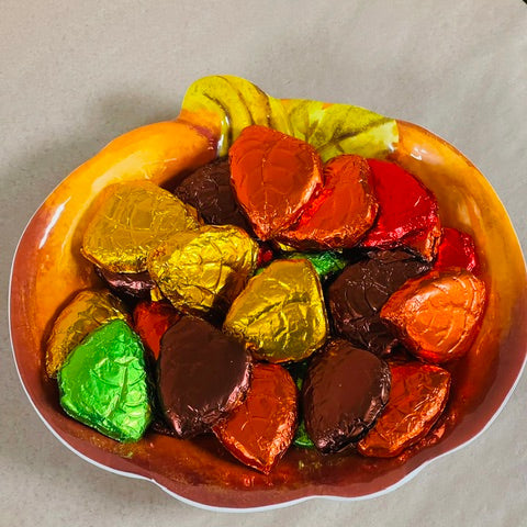 A bowl filled with assorted heart-shaped chocolates wrapped in colorful foil, including gold, orange, red, brown, green, and pink, set on a light beige surface.