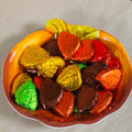 A bowl filled with assorted heart-shaped chocolates wrapped in colorful foil, including gold, orange, red, brown, green, and pink, set on a light beige surface.